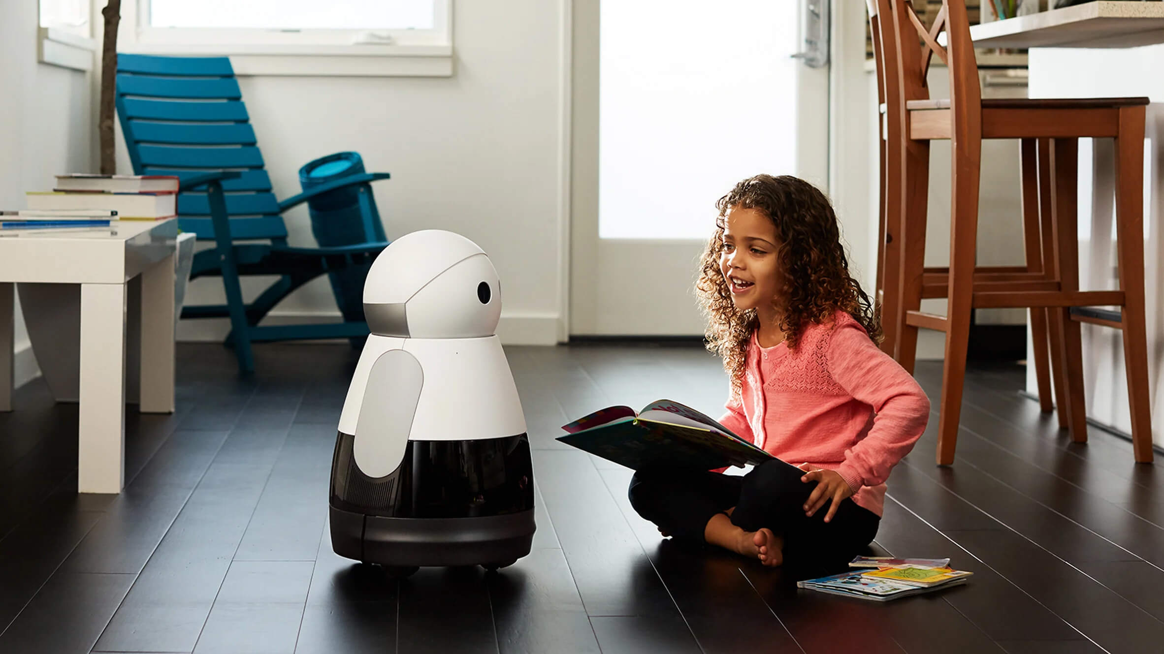 A young girl sitting on the floor reading a book to Kuri the robot companion in a modern living space.