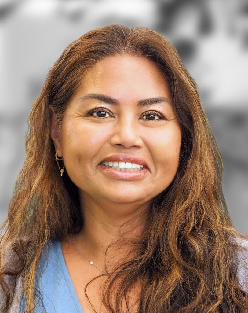 A woman with long, wavy brown hair smiles at the camera. She is wearing a light blue top and gold earrings against a blurred background.