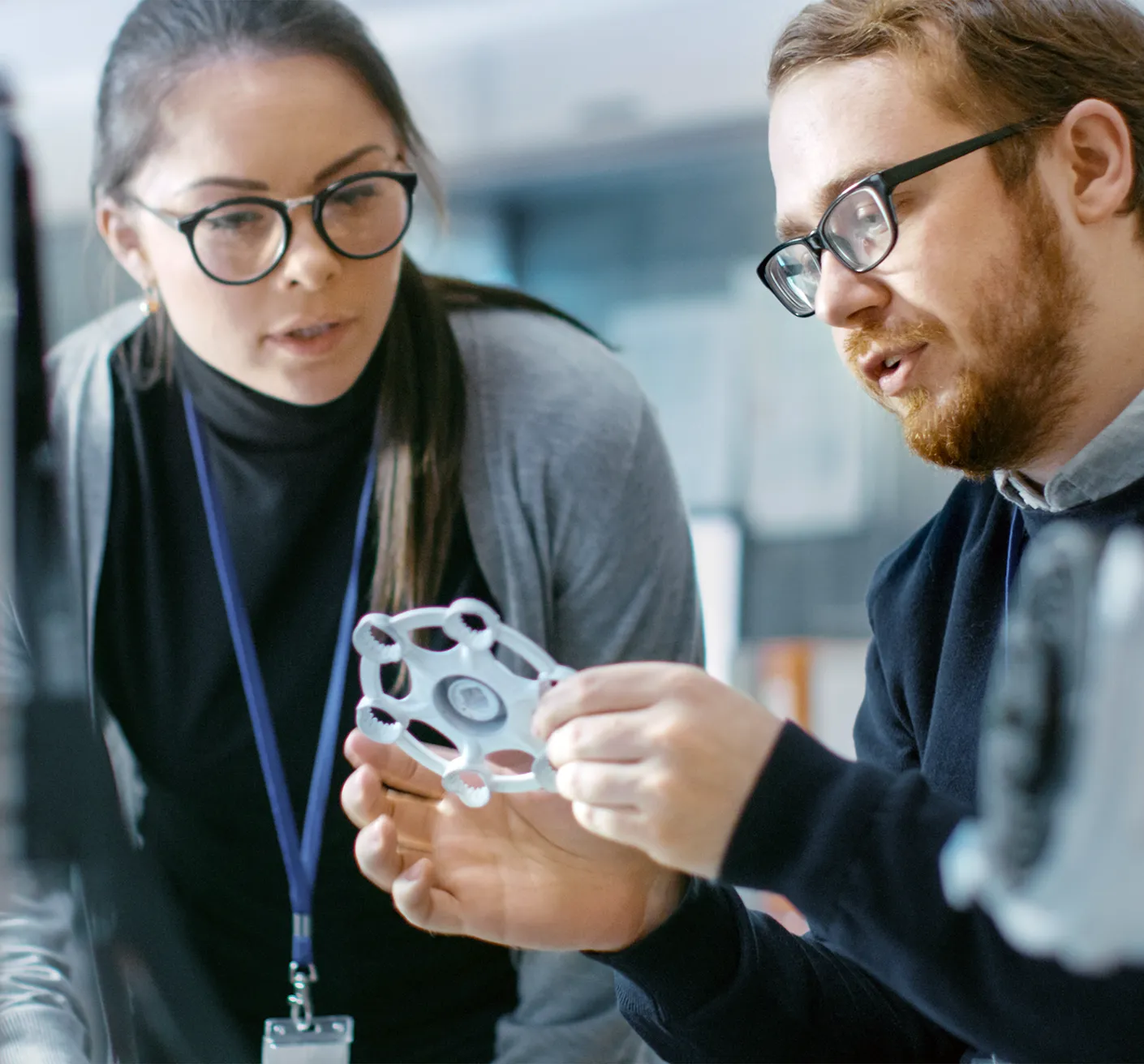 Two people examining a mechanical part in a lab setting. The man holds the part while the woman looks on attentively. They are both wearing glasses and have ID lanyards.