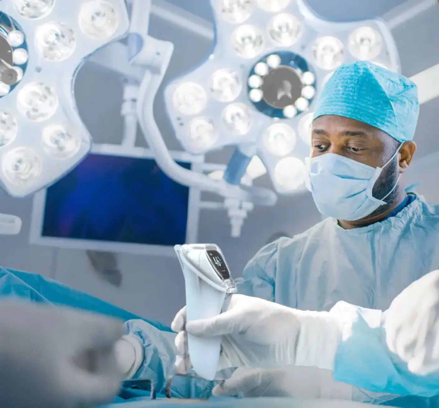 A surgeon in scrubs operates, holding a medical device in an illuminated operating room.