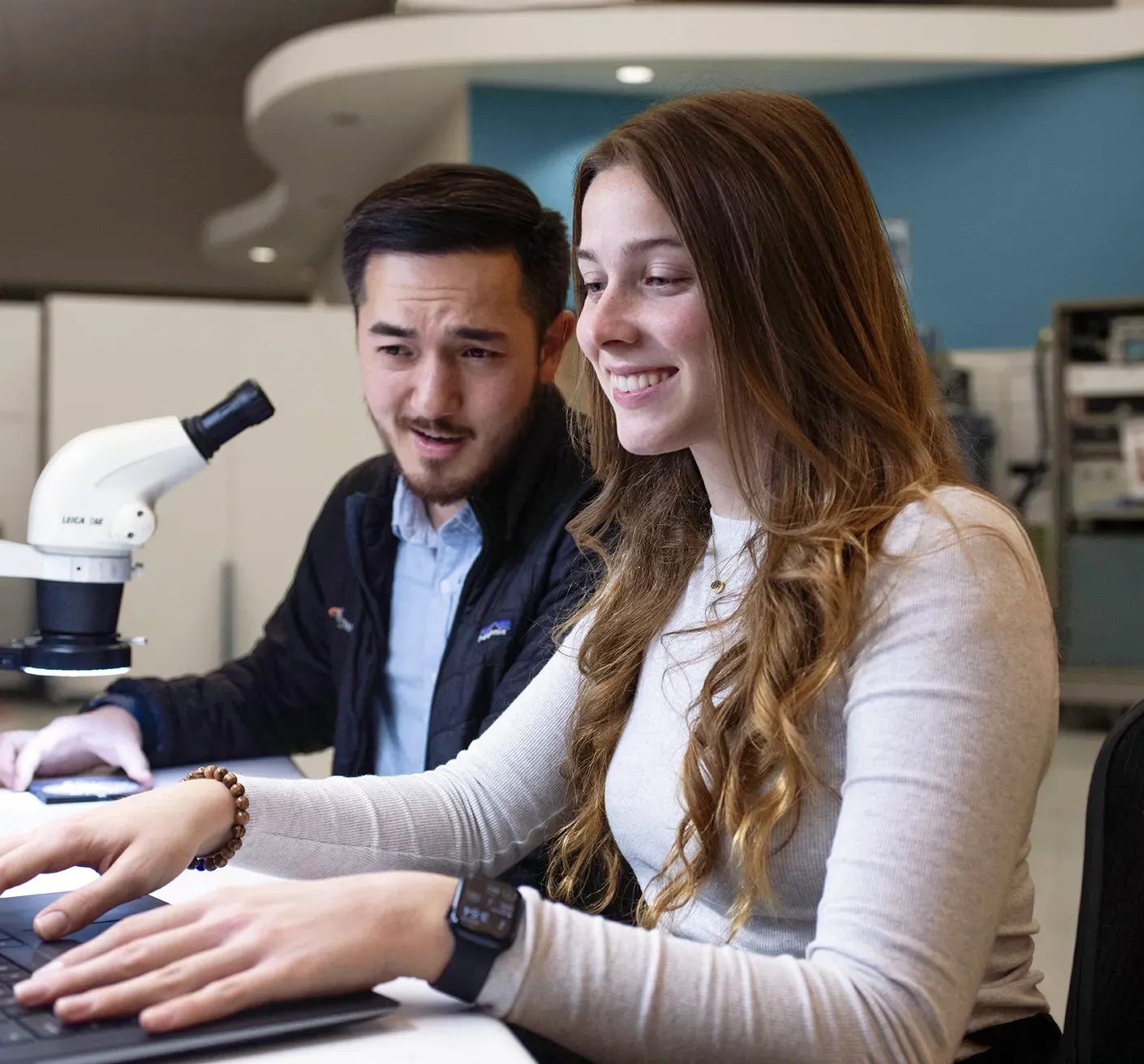 Two people work together at a desk with a laptop and a microscope, focused and collaborating on product development.