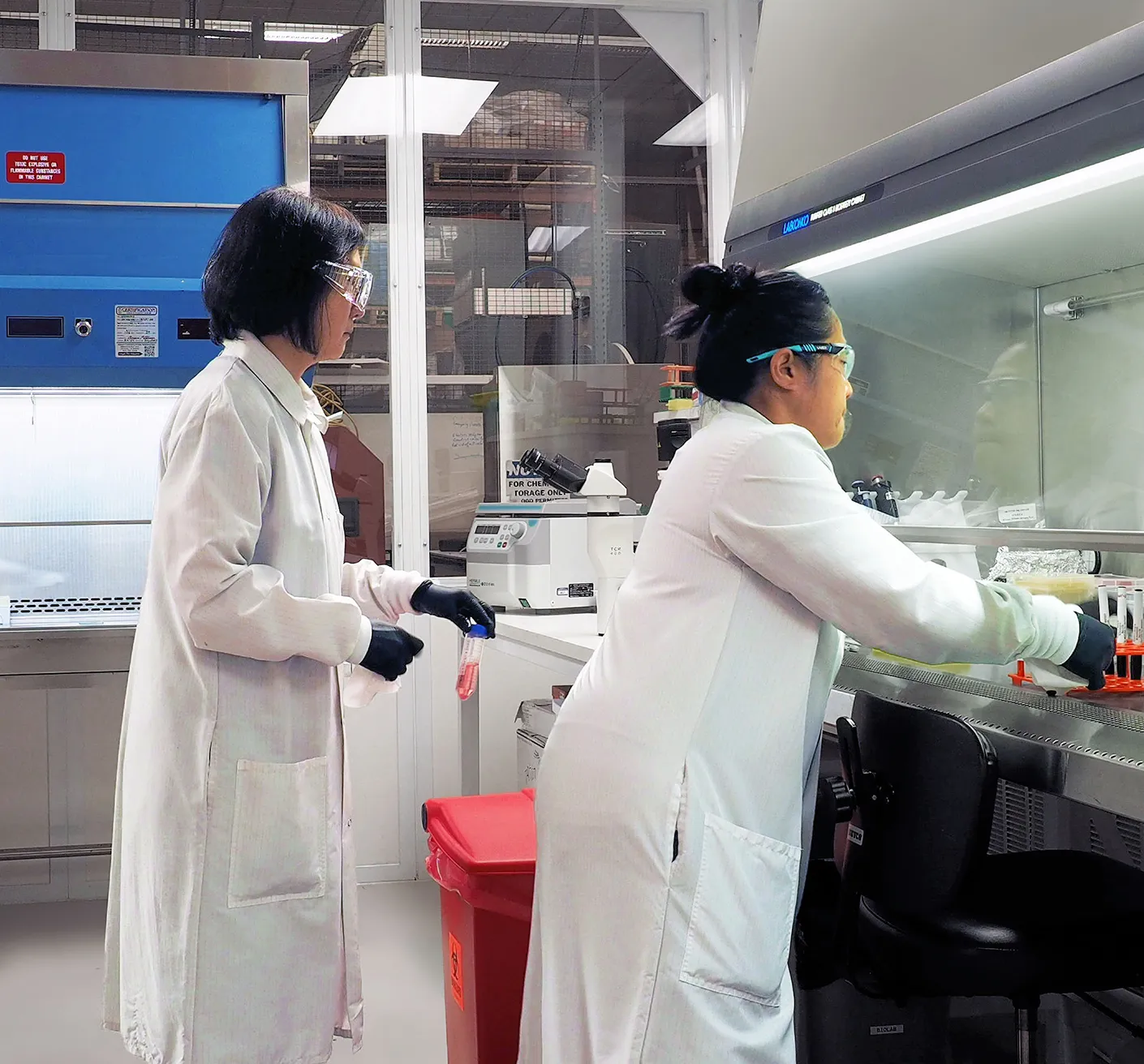 Two Triple Ring scientists in lab coats work in a BSL2 laboratory. One operates equipment at a workstation, and the other holds a small container. Safety gear and lab tools are visible around them.